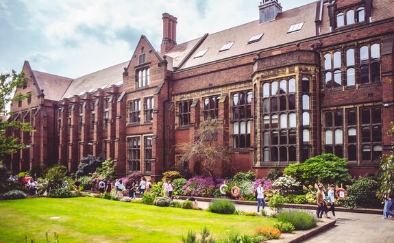 The Armstrong Building on Newcastle University campus with ornate details, a garden in front, and people walking under a partly cloudy sky.